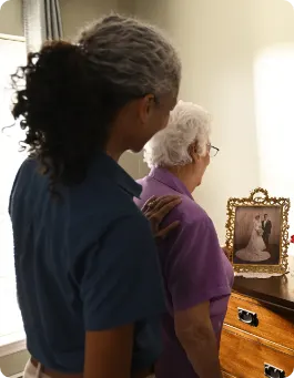 An older female stands facing a dresser looking at a photo. A female care giver stands behind her with a hand on the older female's shoulder.