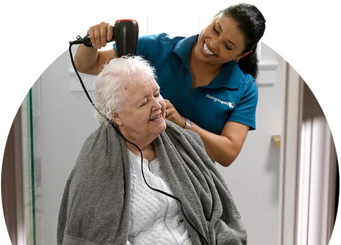An older female sits in a chair with a towel draped across her shoulders. A female care giver stands behind her with a hair dryer, helping the older female dry her hair.