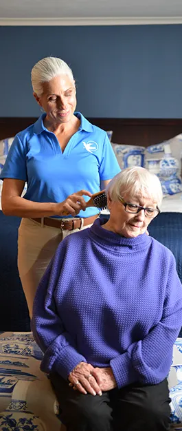 An older woman in a purple blouse sits in a chair while a female care giver stands behind her with a brush in her hand.