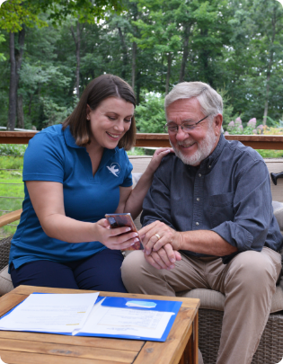 A female Visiting Angels caregiver sits outdoors with a smiling elderly man, showing him something on a smartphone, with documents on the table, highlighting a supportive and engaging respite care environment.