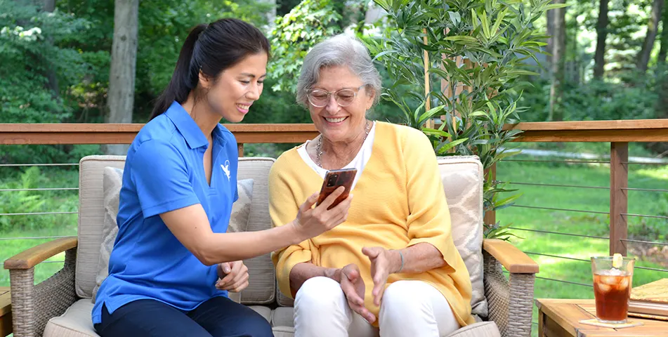 A female Visiting Angels senior home care provider shows an elderly woman a phone in the yard.