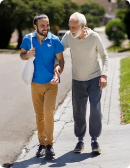 An older man and a male care giver walk on a sidewalk outdoors. Both are smiling. 