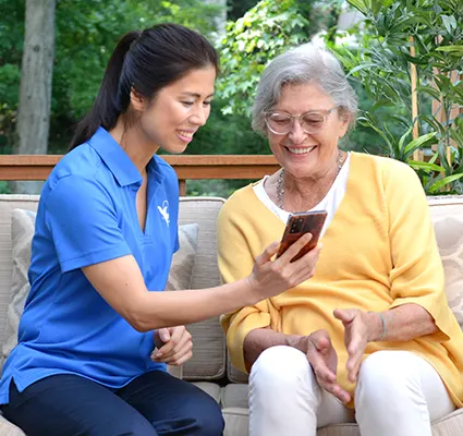 A female Visiting Angels senior home care provider shows an elderly woman a phone in the yard.