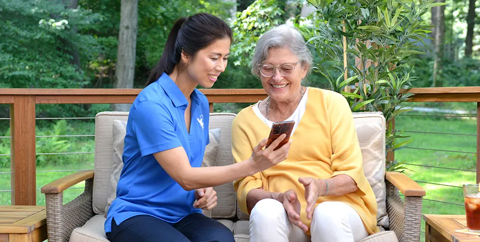 A female Visiting Angels senior home care provider shows an elderly woman a phone in the yard.