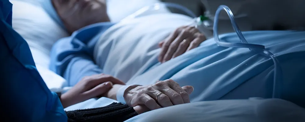 A close up of a patient laying in a hospital bad with the hand of a supporter overlaying theirs. 