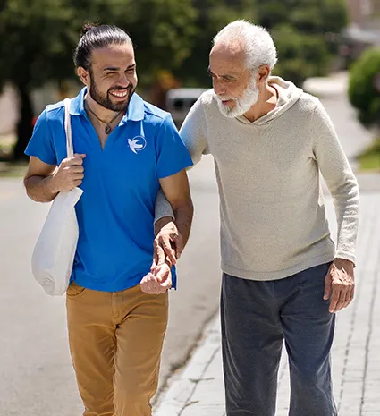 An older man and a younger male care giver walk arm in arm outdoors. Both are smiling. 