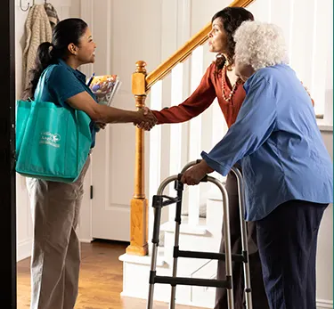 An older female stands next to a staircase with the support of a walker. Another female stnads next to her with her hand outreached to share a female care giver's hand who also stands in the entry way.