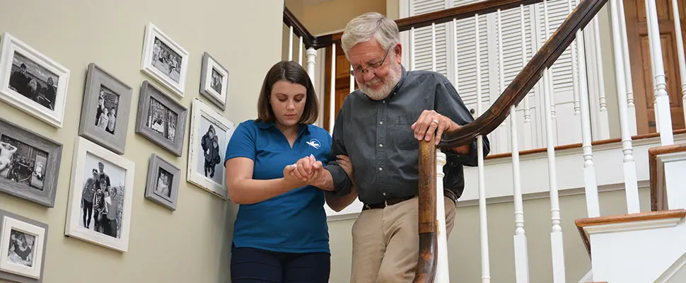 Visiting Angels female caregiver assists an older man walking safely down stairs in his home, providing personalized senior home care and mobility support..