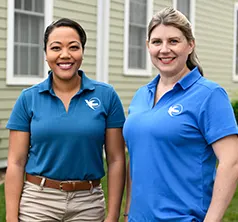 Two female care givers stand side by side, outdoors, and smile at the camera. 