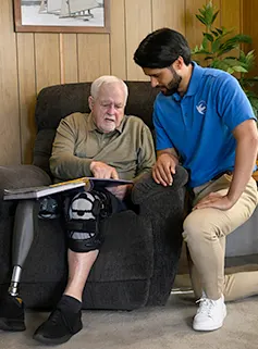 An older man sits in an arm chair looking at a book while a young male care giver kneels next to him. 
