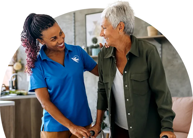 An older woman in a green button down blouse stands with the support of a walker. A female care giver stands next to her, smiling, with a hand on the older woman's shoulder.