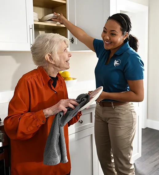 An older woman in an orange blouse dries off a dish with a towel while a female care giver stands close by, putting away another dish.