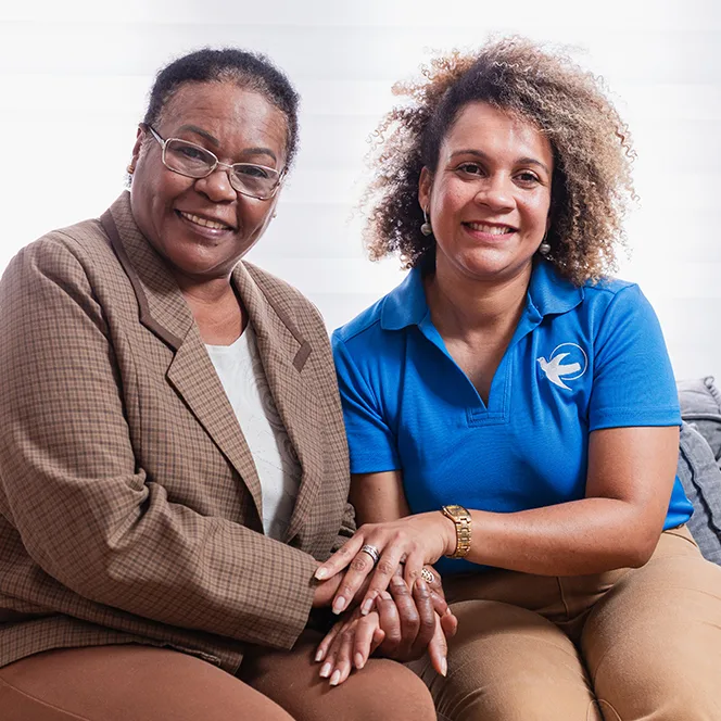 A female care giver sits on a couch next to an older woman. Their hands are intertwined.