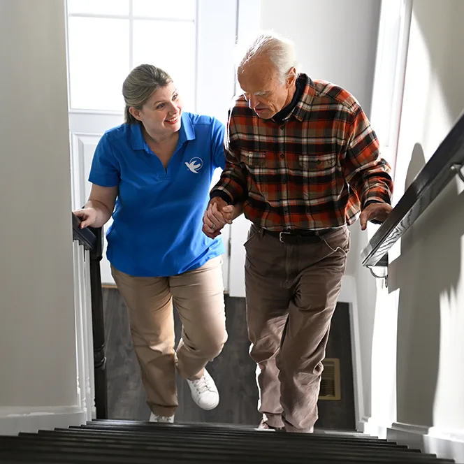 An older man in a plaid button down walks up a set of stairs with his left hand on the railing. A female care giver stands on his right to offer support.