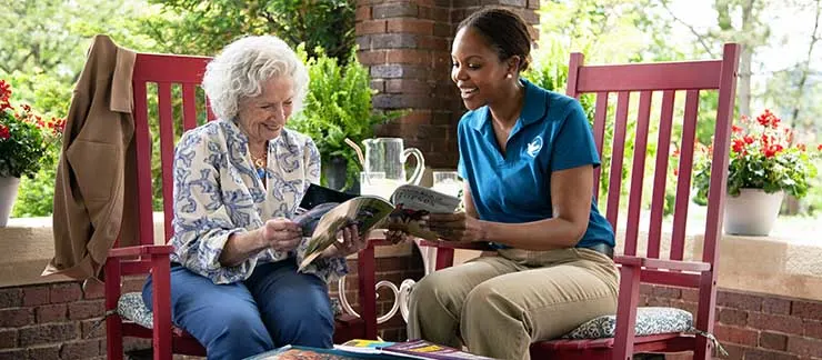 Senior woman reading a magazine on porch with a professional caregiver while aging in place at home.