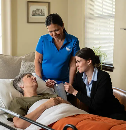A female caregiver and family member comfort an older woman resting in a hospital bed at home.