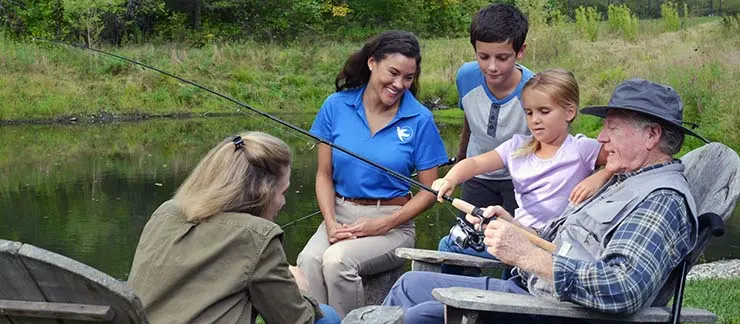 Senior man fishing by a pond with family members and a Visiting Angels caregiver.