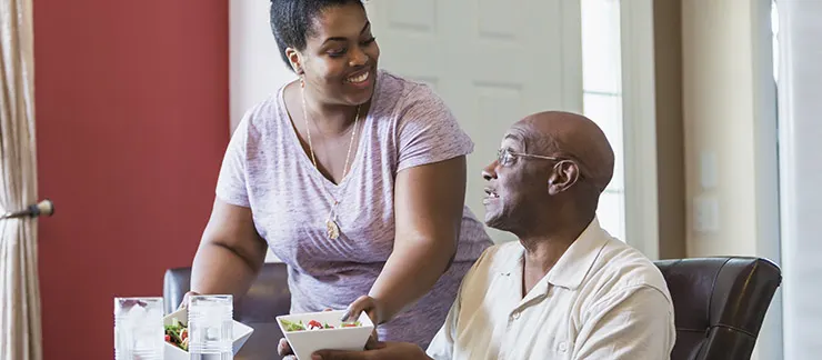 Female home care worker smiles while serving lunch to senior male sitting at table.