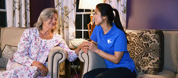 Senior woman living with dementia smiling and holding hands with a Visiting Angels female caregiver at home.