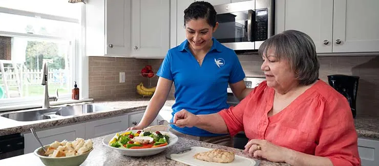 Female in-home caregiver helps a senior woman prepare a healthy meal with fresh vegetables and grilled chicken in her kitchen.