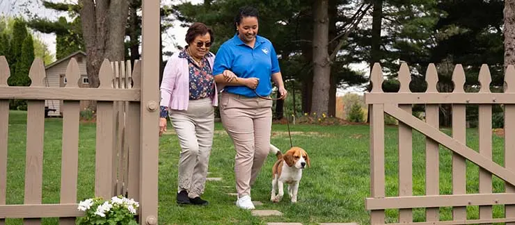 Female caregiver walking with a senior woman and her dog outdoors to promote safe walking and healthy aging