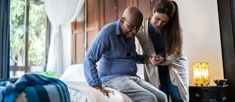 A caregiver helping an elderly man get up from his bed.